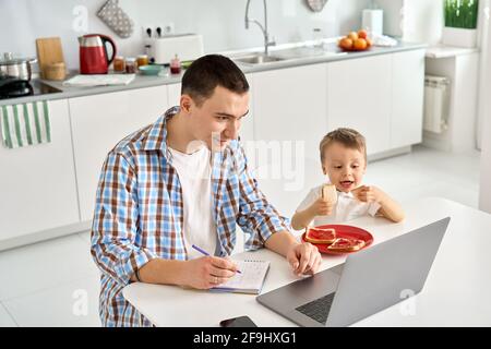 Father working from home office using laptop sitting at table with cute kid son.