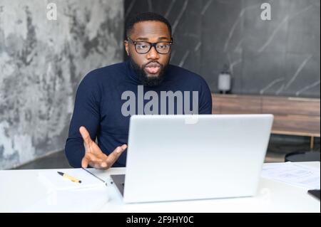 African man in elegant glasses sitting at desk working at laptop in the home office, young entrepreneur explains to his team the tasks for a week or a promotion strategy using a video conference Stock Photo