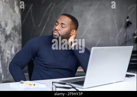 Exhausted African American worker felt asleep at workplace sitting at the laptop, overworked, Feeling exhausted, frustrated young beard man holding the back of the head, professional burnout syndrome. Stock Photo