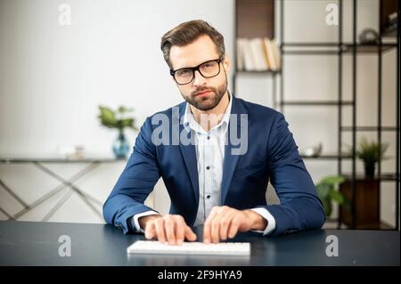 Serious elegant business man sitting in the modern spacious office typing on the white keyboard, looking directly at camera, chatting with business partners, managing large company remotely, big boss Stock Photo