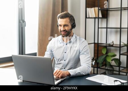 Cheerful mature fair-haired man freelancer in headset working from home during pandemic, having video chat with employer or clients, using modern laptop and documents, copy space, typing on keyboard Stock Photo