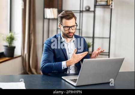 Slavic stylish man talking at laptop by video call, focused young businessman in eyeglasses involved in online video call negotiations meeting with partners colleagues or studying distantly at home Stock Photo