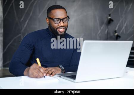 Young african man in elegant glasses searching information for project using laptop wifi connection indoors, male freelancer writing article in textbook for add interesting content on website web page Stock Photo
