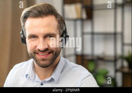 Head shot European man wear headset on blurred background with shelves and leaves, looks calm, joyful and peaceful, using videocall conferencing app, company consultant, teacher portrait, easy e-learn Stock Photo