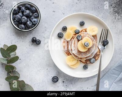 pancakes with fresh blueberries, banana in white plate on gray background with branch of eucalyptus. healthy food concept. horizontal image, top view Stock Photo
