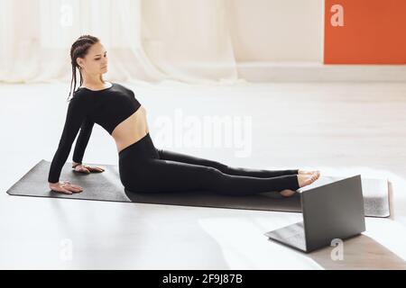 Woman in black sportswear, practicing yoga, working out in the studio sitting on the mat and looking closely at the laptop screen, doing online exerci Stock Photo