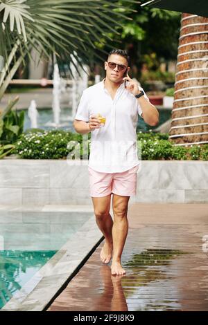 Handosme man with glass of bourbon walking by swimming pool and talking on phone with business partner Stock Photo