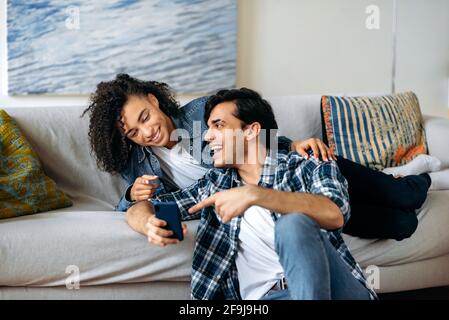 A loving young multiracial couple spends leisure time at home on the sofa, using a smartphone, browsing the Internet and social networks, laughing, the guy shows the girl something funny on the phone Stock Photo