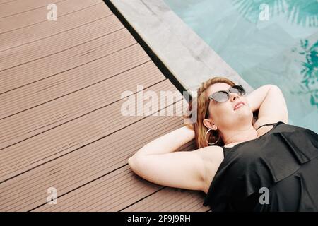 Smiling mature woman in sunglasses sunbathing by swimming pool, view from above Stock Photo