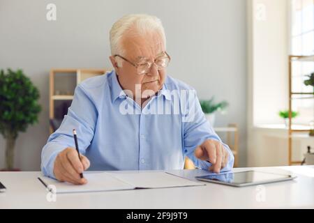 Elderly man worker finding information in internet on tablet and writing down in notebook Stock Photo