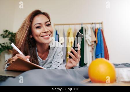 Portrait of smiling attractive young woman lying on bed, checking her phone and taking notes in planner Stock Photo