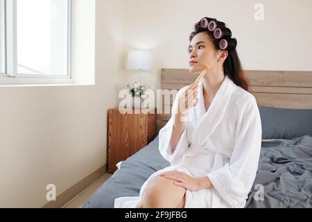 Attractive young Vietnamese woman in bathrobe massaging face with roller after taking morning shower Stock Photo