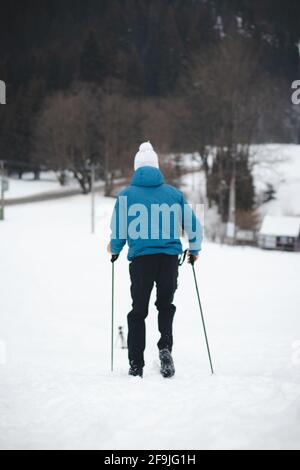 young man wearing blue jacket and white cap runs down hill using trekking sticks for stability and better descent with no falls. Winter mountain runni Stock Photo