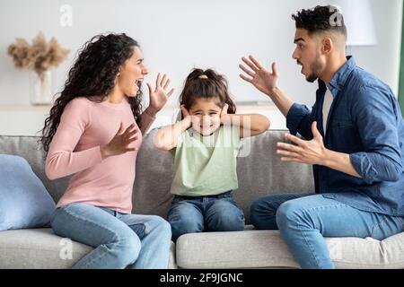 Parental Conflicts. Upset Little Girl Covering Ears Not To Listen Parents Arguing Stock Photo