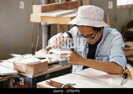 Concentrated young Asian man using chisel and hammer when working with wood board Stock Photo