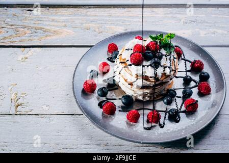 colorful plate of pancakes with cream, mint leaves and fresh berries with chocolate syrup falling on top on wooden table Stock Photo