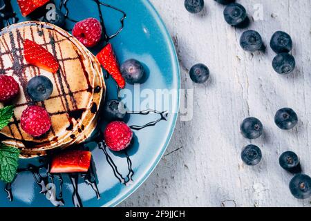 top view of delicious pancakes with chocolate syrup, fresh fruits and mint leaves on a blue plate on wooden table. blueberries background. copy space Stock Photo