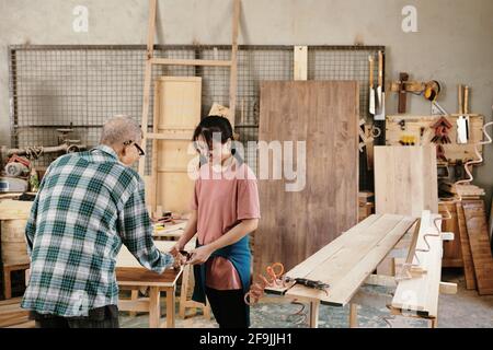Senior Asian carpenter showing granddaughter how to cover sanded wood with oil, family business concept Stock Photo