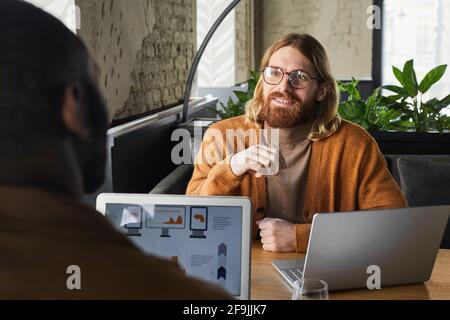 Portrait of contemporary bearded businessman smiling at partner during meeting at table in cafe, copy space Stock Photo