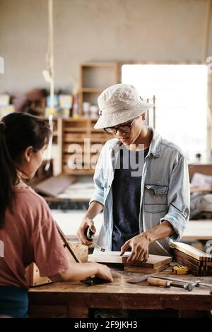 Professional young Asian carpenter measuring wooden board with tape measure when working on project at workbench Stock Photo