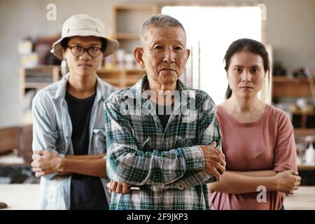 Team of senior experienced carpenter and his young apprentices standing workshop with arms folded and looking at camera Stock Photo