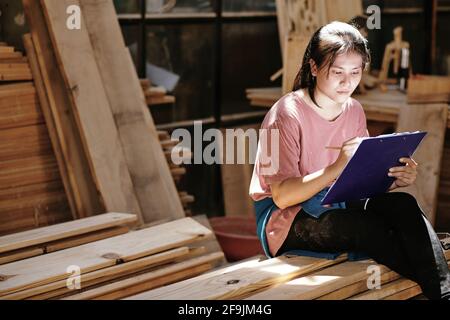 Creative young female customer sitting on pile of wooden boards and drawing furniture item blueprint Stock Photo