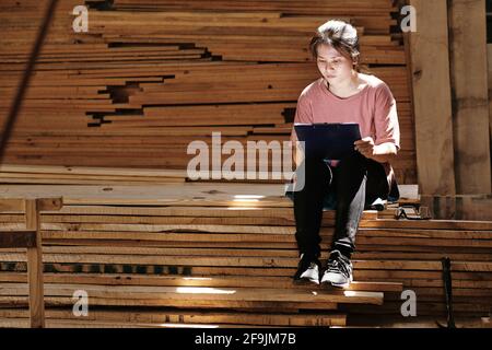 Talented creative young woman sitting on pile of wooden boards and drawing sketch of furniture item for customer Stock Photo