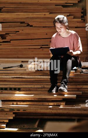 Young female carpenter working on design of furniture item for customer and drawing sketch she sitting on pile of wooden planks Stock Photo