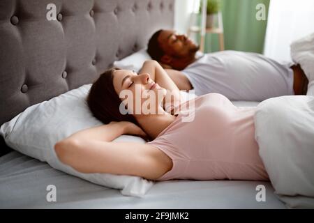 Happy married interracial couple resting in bed at home, lying with hands behind their heads in morning, feeling relaxed Stock Photo