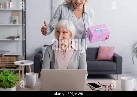 Smiling woman holding present near elderly mother using laptop Stock Photo