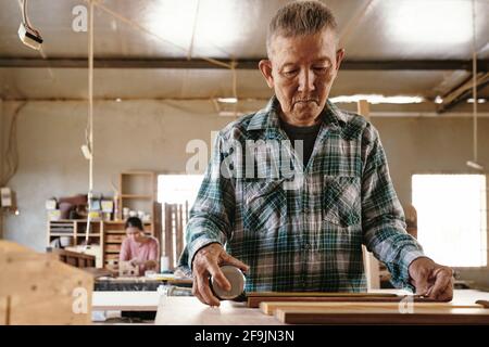 Concentrated senior carpenter measuring wooden planks with tape measure on his workbench Stock Photo