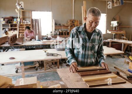 Serious senior Vietnamese carpenter putting sanded and oiled workpieces in a raw on workbench Stock Photo