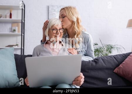 Adult woman kissing smiling mother with laptop on blurred foreground Stock Photo