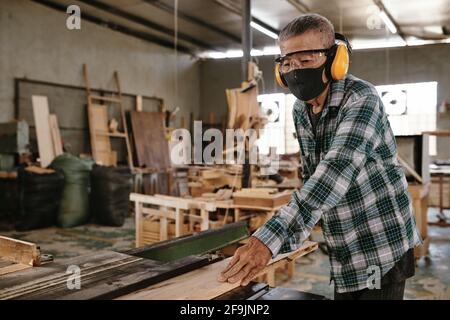 Senior man in protective mask and earmuffs working with wood in carpenters workshop Stock Photo
