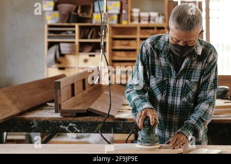 Elderly experienced carpenter in protective mask polising wooden plank with electric sander Stock Photo