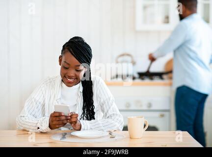 Young African Lady Using Smartphone In Kitchen While Her Husband Cooking Lunch Stock Photo