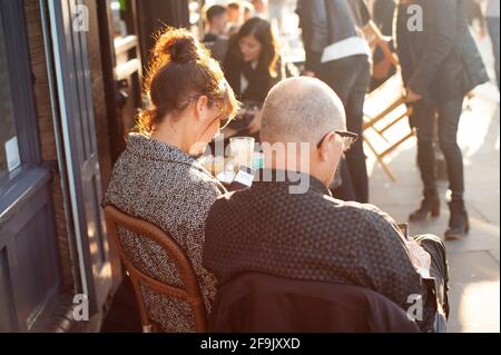 Rear view of disconnected middle aged British couple, checking their mobile phones outside a coffee shop. Broadway Market, E2, East London. Nov 2015 Stock Photo