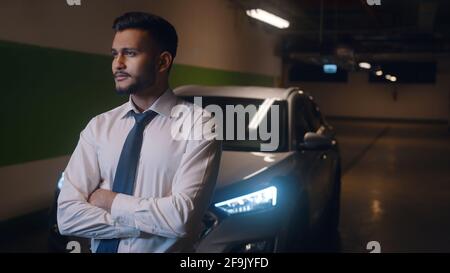 Waist up of adult businessman standing indoors in front of his car with arms crossed and looking ahead. Stock Photo