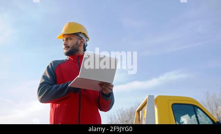Waist up low angle view of construction engineer with safety helmet standing on-site with laptop. Blue sky with soft clouds in background. Stock Photo