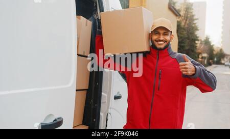 Smiling adult delivery man carrying cardboard box, showing thumb up gesture and looking at camera. Parcel delivery from eshop concept. Stock Photo