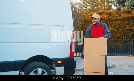Smiling mid adult delivery man outside white van standing at stacked blank boxes. Parcel delivery concept. Stock Photo