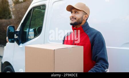 Smiling bearded man wearing beige cap looking away. Delivery man waiting with big cardboard parcel at car. Stock Photo