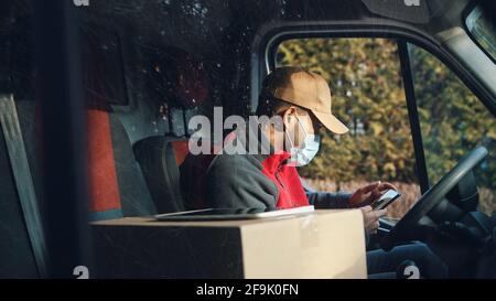 Employer of parcel delivery service sitting in car and using mobile phone. View from passenger side. Stock Photo
