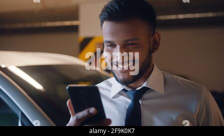 Adult mixed race man using mobile phone indoors. Smiling businessman making video call in underground garage. Stock Photo