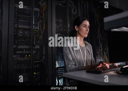 Portrait of female server engineer using laptop while sitting in dark IT room, copy space Stock Photo