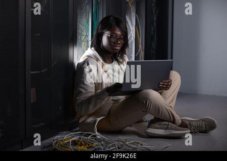 Full length portrait of young African-American woman using laptop in server room and sitting on floor while working with supercomputer at data center, Stock Photo