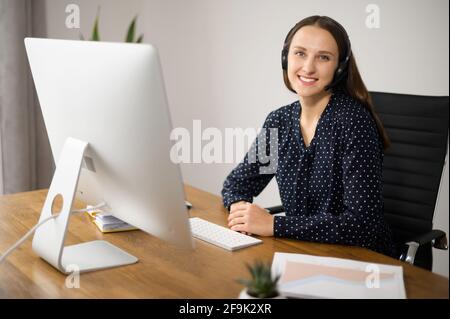 Smiling young caucasian woman wearing wireless headset sits at the workplace in front of PC, looks at the camera and smiles. Friendly female support center operator Stock Photo