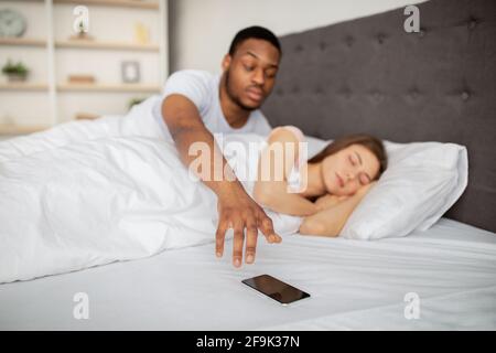 Suspicious black husband checking his sleeping wife's cellphone in bed at home. Relationship problems Stock Photo