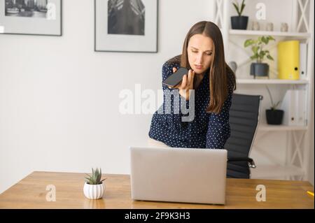 Busy female employee is using voice assistant on the smartphone, young woman talking in the mic of her phone, recording voice message, using voice recognizable function or app and typing on the laptop Stock Photo