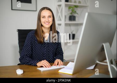 Young smiling woman is using PC sitting at the desk in home office, female employee typing and looks at the camera Stock Photo